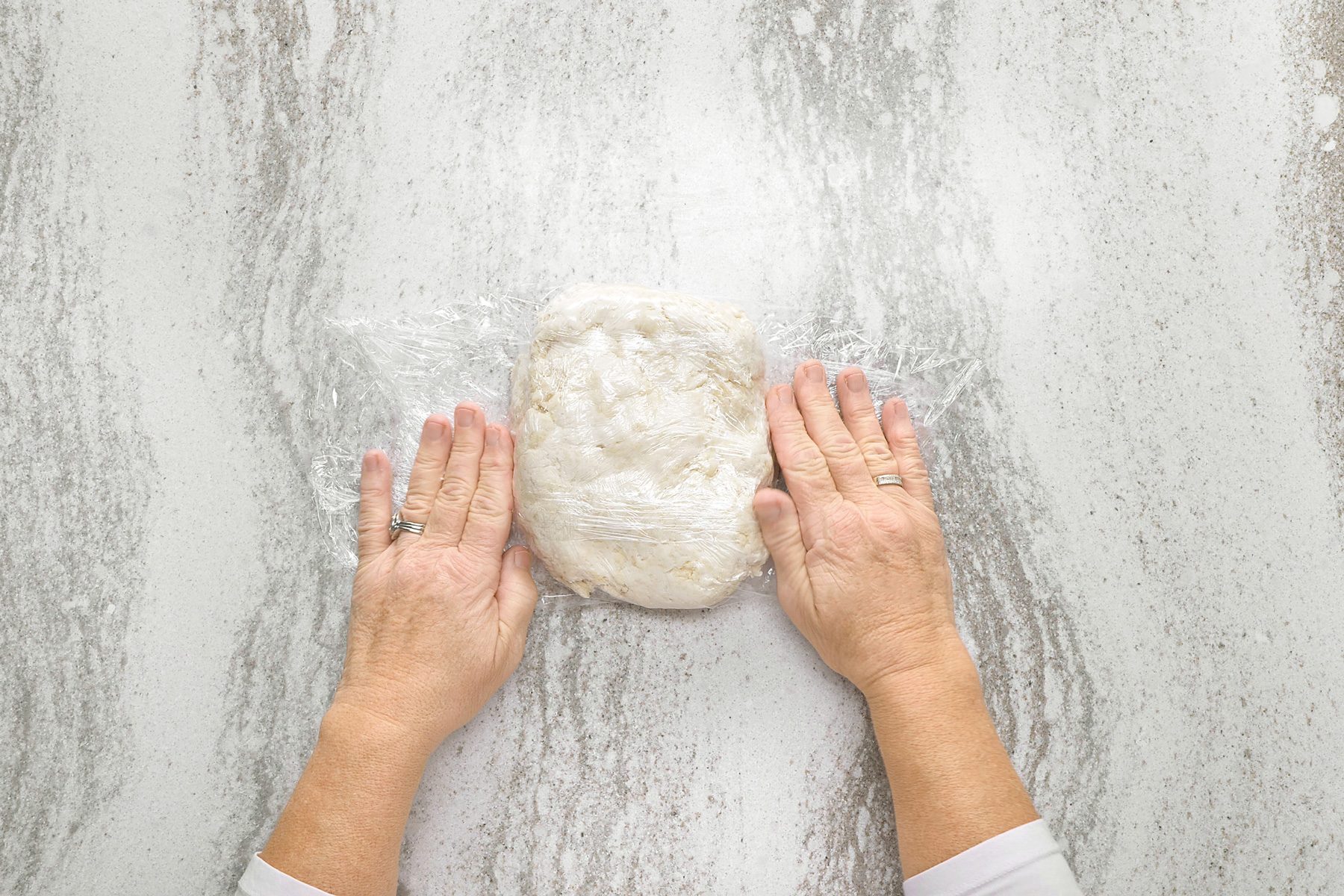 overhead shot of a person's hands wrapping a ball of dough in plastic wrap, the dough is a light color; the hands are on a white textured surface