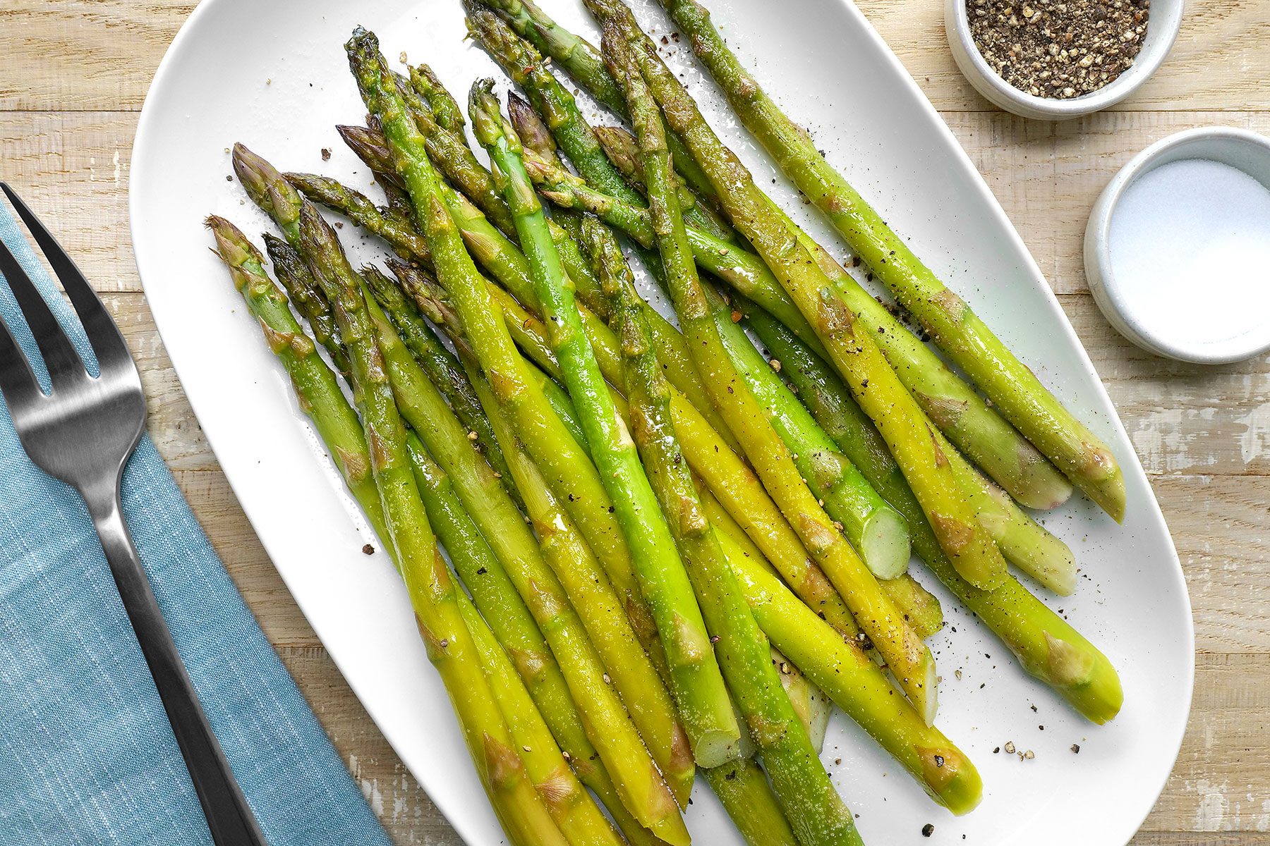 A plate of seasoned and roasted asparagus spears on a white oval dish, garnished with cracked black pepper. A fork rests on a blue napkin beside the plate, and small bowls of salt and pepper are nearby.