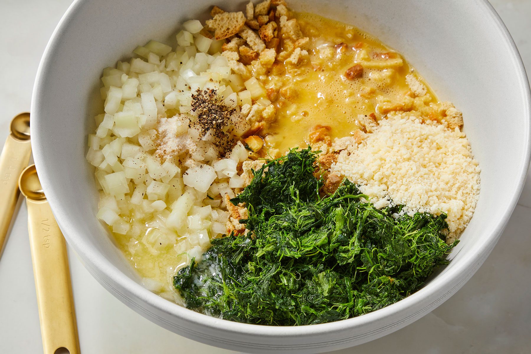 A large white bowl containing chopped onions, bread crumbs, a mixture of beaten eggs and seasonings, grated cheese, and chopped fresh herbs. Two gold measuring spoons are placed beside the bowl on a light surface.