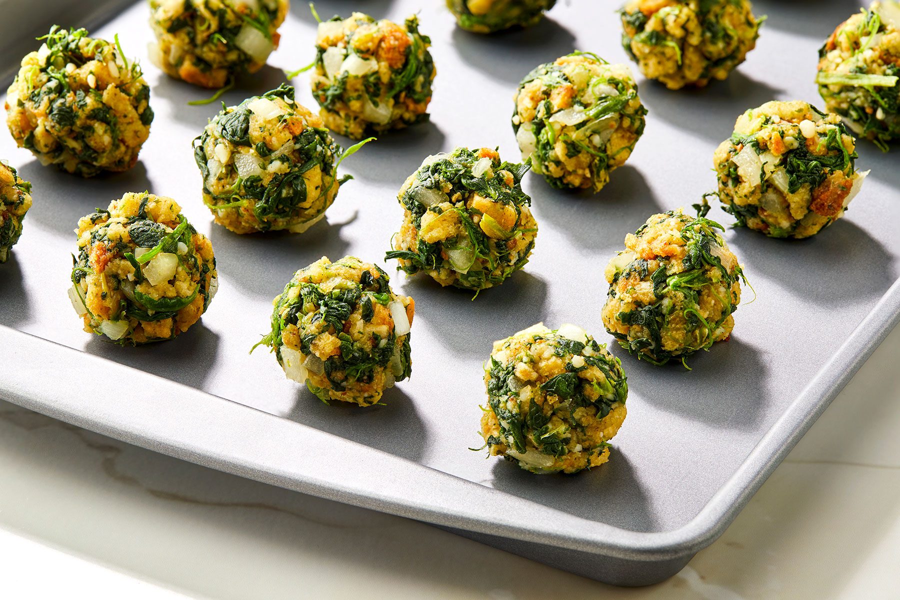 A baking tray holds evenly spaced spinach balls, featuring a mix of chopped spinach, herbs, and visible chunks of nuts. The balls are evenly placed on the metal tray, ready for cooking or baking.