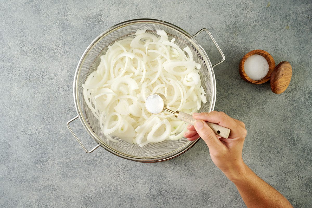 Overhead view of sweet onions in a colander being sprinkled with canning salt and left to drain over a plate for the Taste of Home Pickled Sweet Onions recipe.