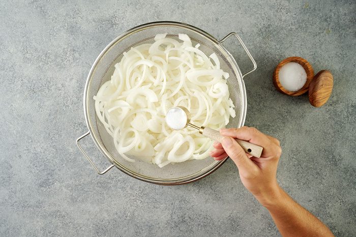 Overhead view of sweet onions in a colander being sprinkled with canning salt and left to drain over a plate for the Taste of Home Pickled Sweet Onions recipe.