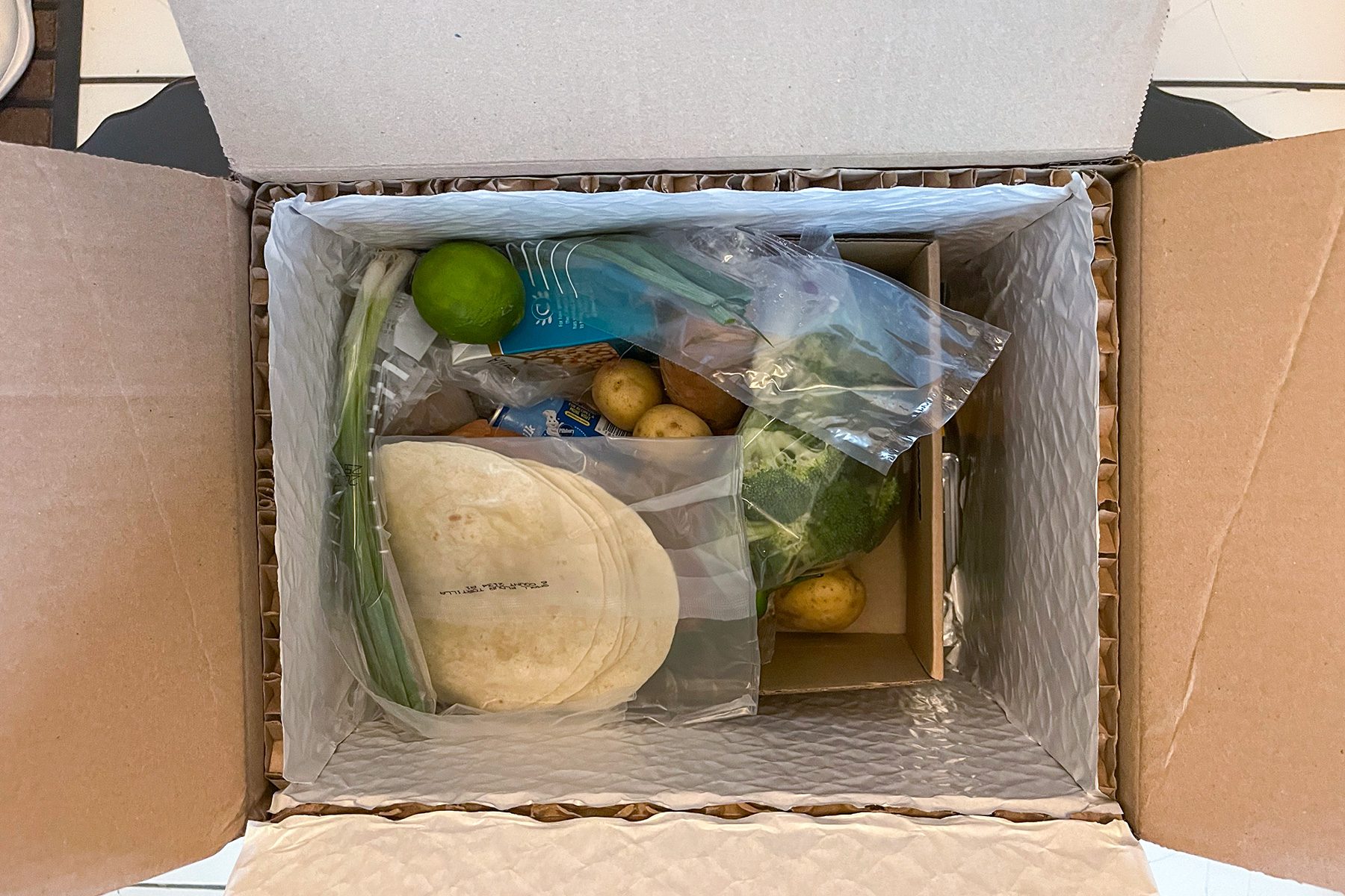 An open cardboard box containing various fresh food items, including potatoes, a lime, tortillas, green onions, and other vegetables in clear plastic bags.
