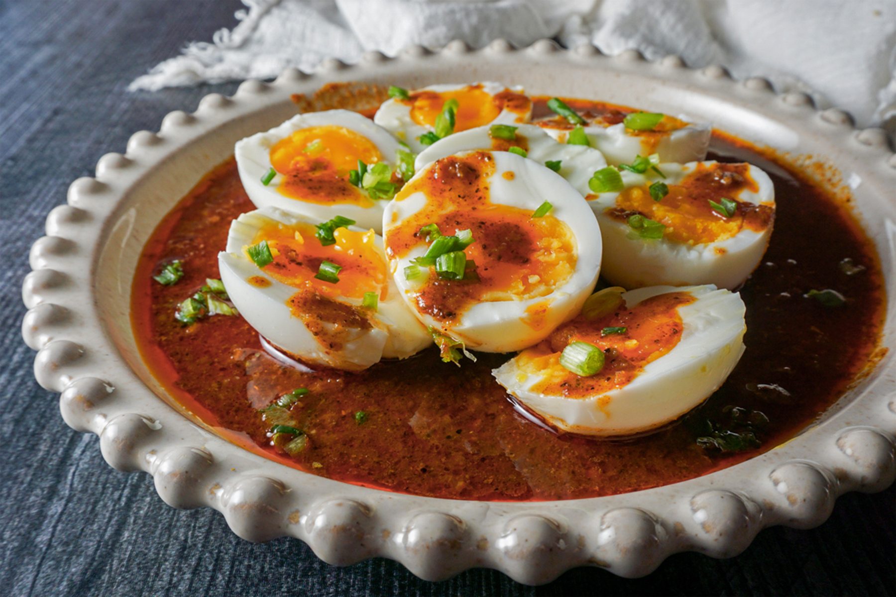 A plate of halved boiled eggs topped with chopped green onions and a spicy red sauce, served on a decorative ceramic dish.