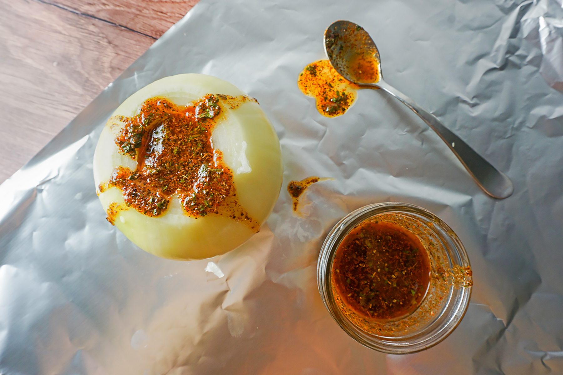 A round cheese wheel with a spicy seasoning mix on top sits on aluminum foil. A small glass jar next to it contains more of the seasoning. A spoon with some of the mix rests on the foil. The table surface is wooden.