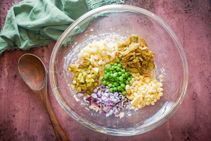 A clear glass bowl on a pink surface contains chopped ingredients: onions, canned tuna, green peas, corn, and red onions. A wooden spoon lies beside the bowl, with a green cloth in the background.