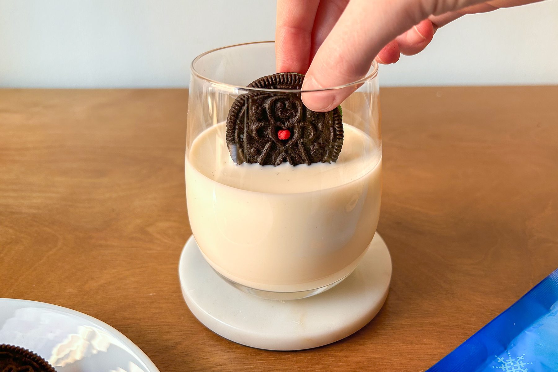 A hand dipping a chocolate sandwich cookie with a red dot into a glass of milk on a wooden table. A small plate with cookie crumbs is visible on the side.