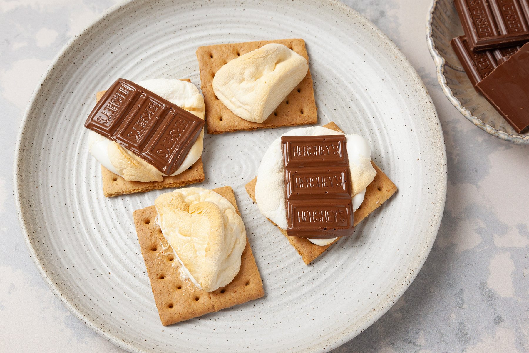 A plate with four s'mores made of graham crackers, toasted marshmallows, and chocolate bars. A bowl with chocolate pieces is next to the plate.
