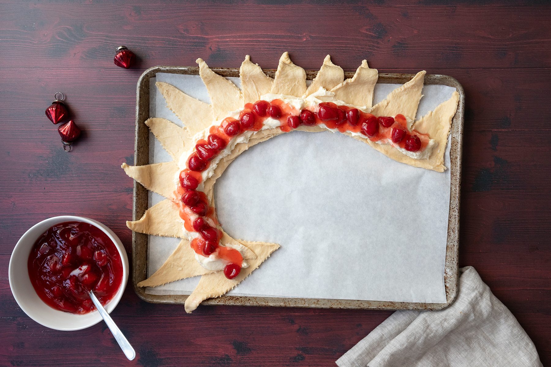 Crescent roll dough shaped like a heart with cherry pie filling and cream cheese on top, arranged on a baking sheet with parchment paper. A bowl of cherry filling and small foil-wrapped chocolates are on the side.