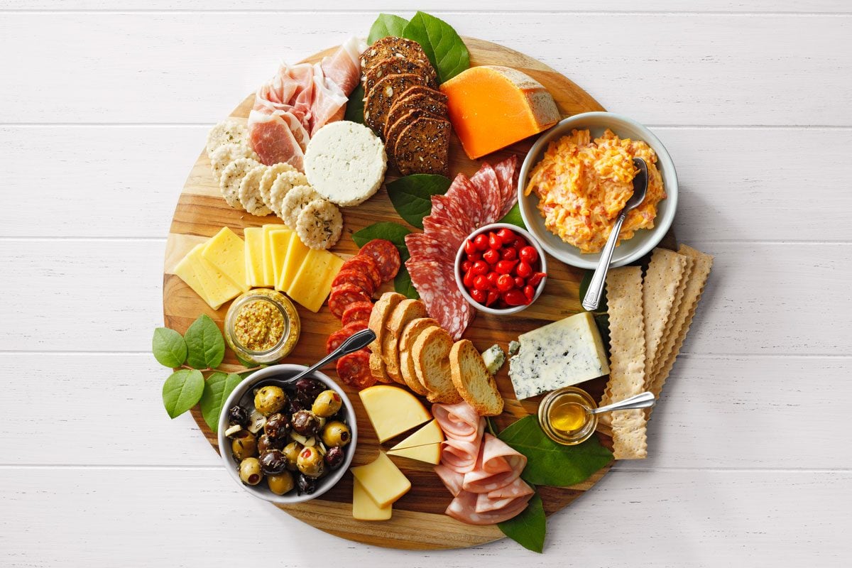A wooden board with a variety of meats, cheeses, crackers, and spreads. Includes sliced salami, prosciutto, cheddar, brie, olives, cherry tomatoes, mustard, and a cheese spread in bowls. Decorated with green leaves.