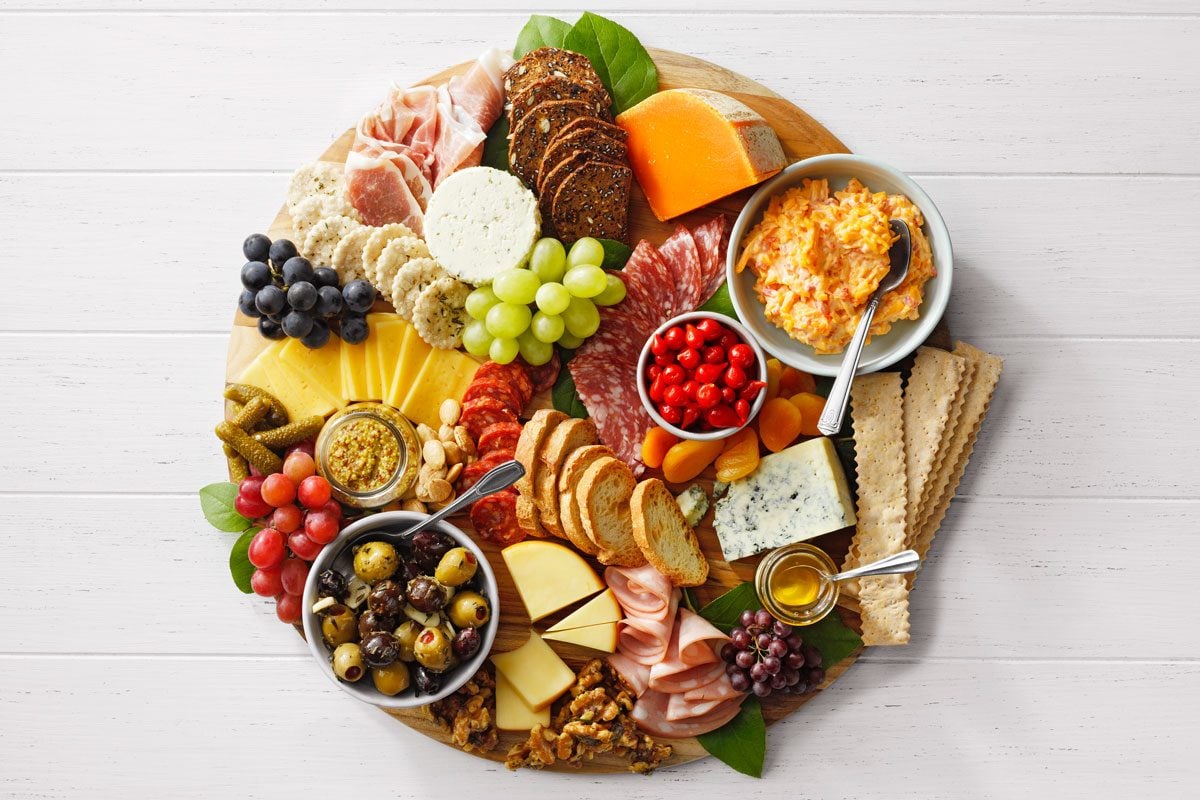 A round wooden board filled with assorted cheeses, meats, crackers, grapes, cherry tomatoes, olives, and nuts. Two small bowls contain spreads. The arrangement creates a colorful and appetizing charcuterie display on a white surface.