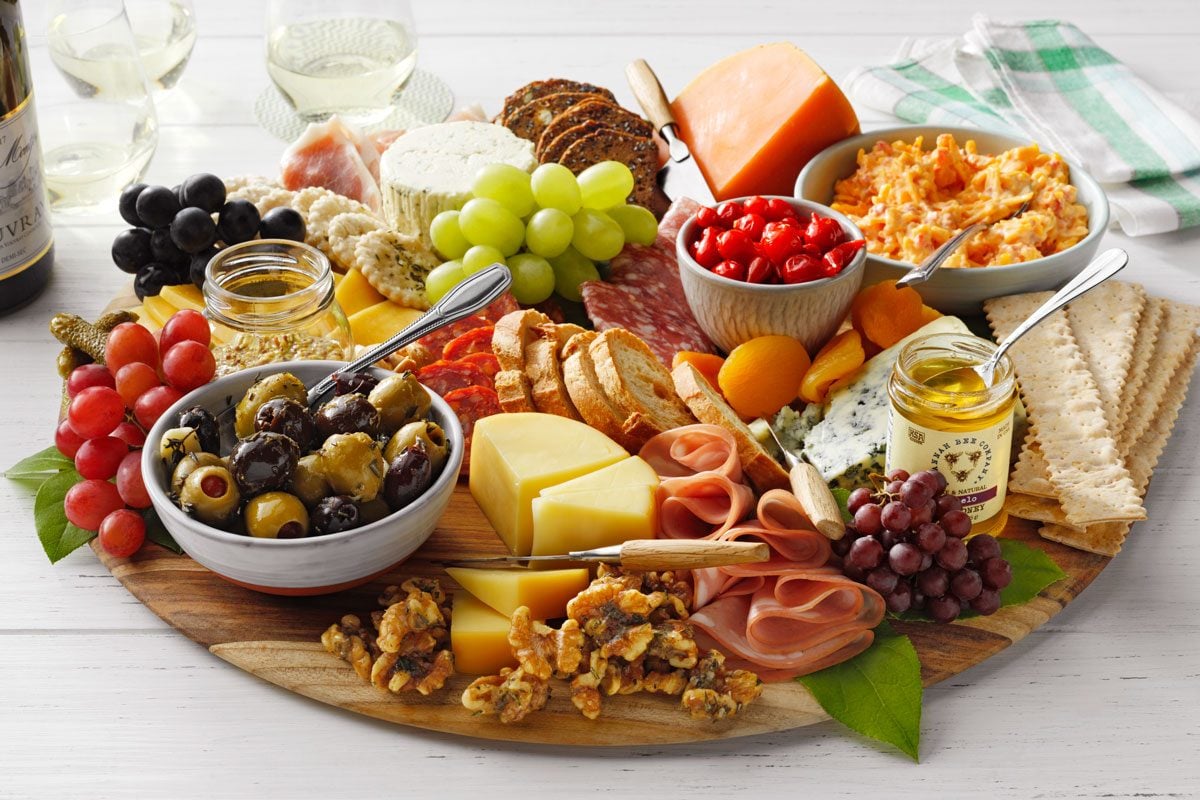 A colorful charcuterie board featuring assorted cheeses, meats, grapes, olives, cherry tomatoes, crackers, nuts, and pomegranate seeds. A glass of white wine and a checkered napkin are in the background.