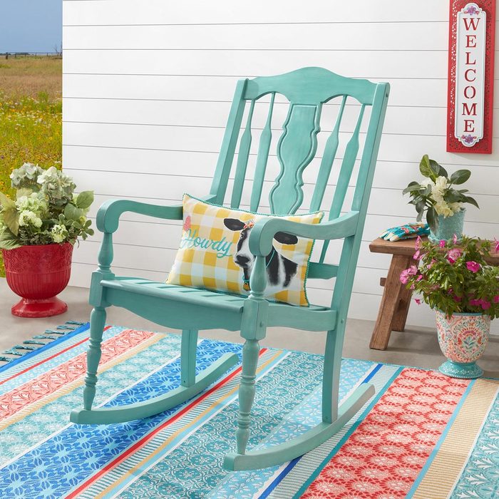 A teal rocking chair with a yellow and white pillow sits on a colorful rug on a porch, with a white wall and plants in the background