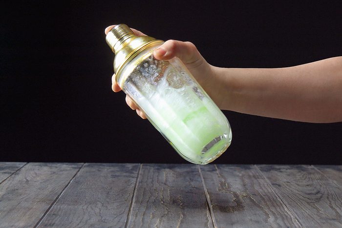 A hand holds a cocktail shaker with a golden top, vigorously shaking it. The shaker is filled with a light green liquid. The background is dark, and the surface is a textured wooden table.