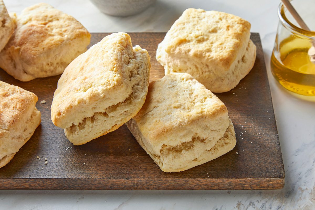 A wooden board with freshly baked biscuits, golden brown and flaky. A jar of honey with a dipper is in the background on a white marble surface. The scene is well-lit, highlighting the biscuits' texture.