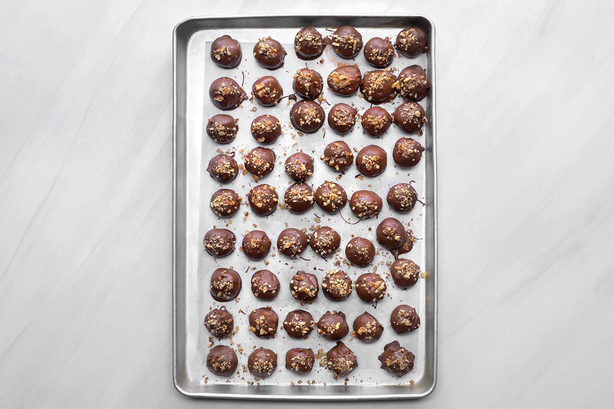 A tray of chocolate-covered treats arranged in neat rows on a parchment-lined baking sheet. The treats are sprinkled with chopped nuts, placed evenly over a light gray marble surface.