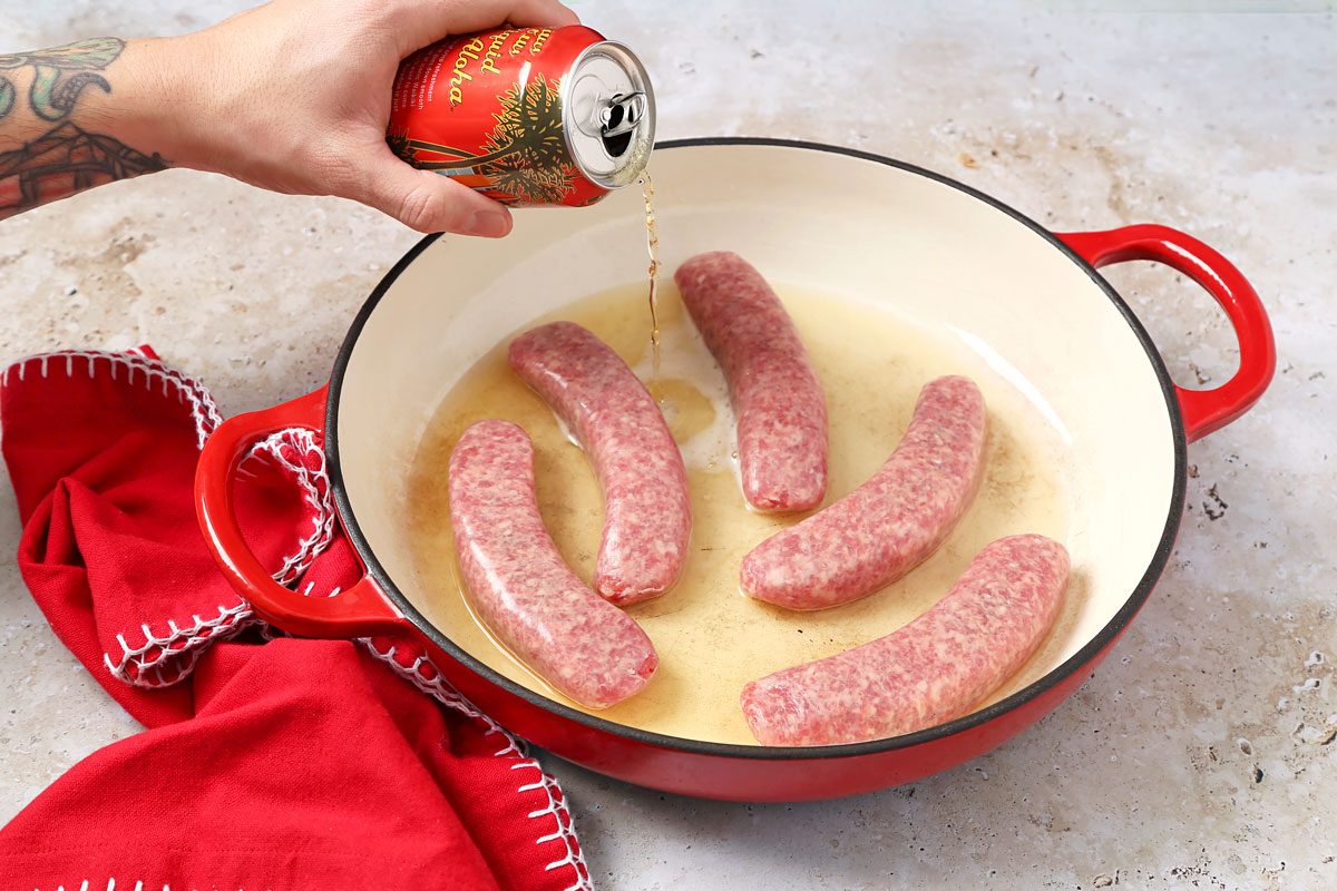 3/4th shot of a person's hand is pouring a can of Aloha beer into a red cast iron skillet that contains five raw sausage links; a red cloth with white crochet trim lays on the counter below the skillet
