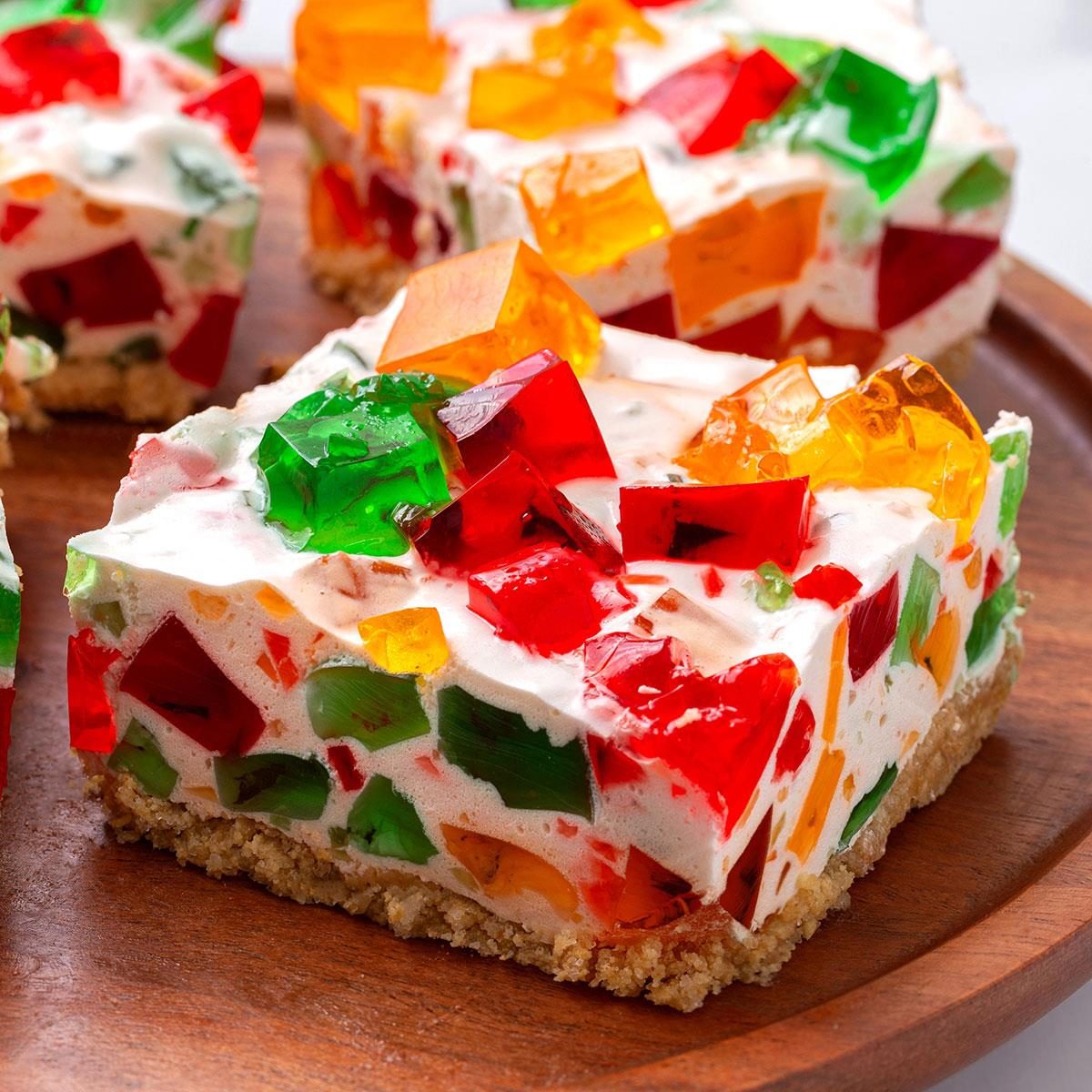 A colorful dessert bar featuring cubes of red, orange, yellow, and green gelatin embedded in creamy filling atop a crumbly crust, presented on a wooden plate.