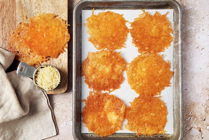 Tray with six golden-brown cheese crisps on parchment paper. A wooden board with a small bowl of grated cheese and a beige cloth is placed beside the tray. The background is a textured off-white surface.