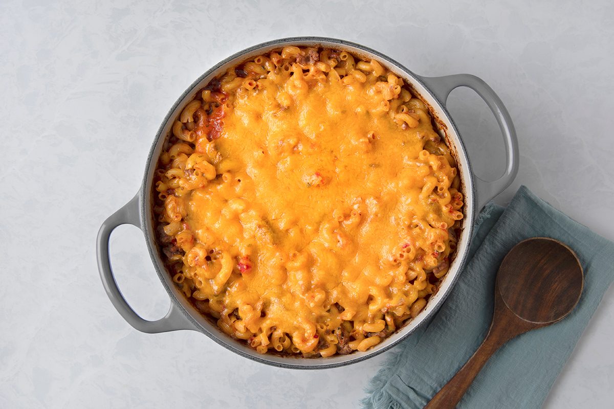 A casserole dish filled with baked macaroni and cheese topped with melted cheddar sits on a blue cloth napkin. A wooden spoon is placed beside it on a light gray surface.