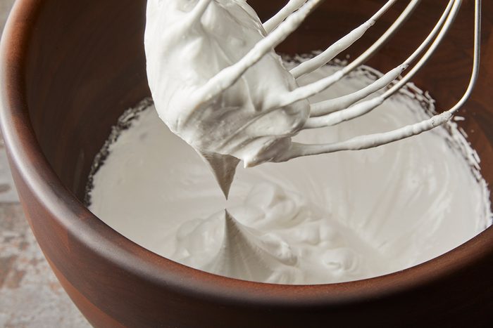 A whisk with creamy white whipped mixture in a wooden bowl, forming soft peaks.