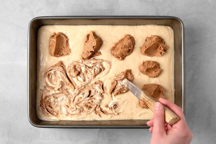 A person's hand using a spatula to swirl brown sugar and cinnamon mix into light-colored batter in a rectangular baking pan, placed on a gray counter.