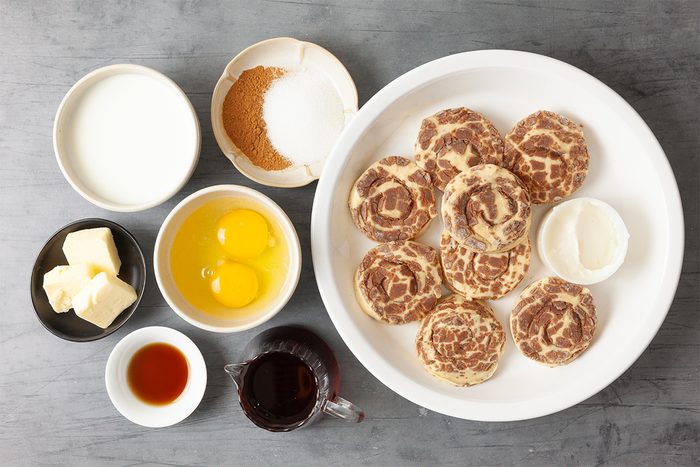 A flat lay of ingredients for cinnamon rolls. A large plate with unbaked rolls, bowls containing eggs, butter, cream, sugar, brown sugar, vanilla extract, and syrup are arranged on a gray surface.