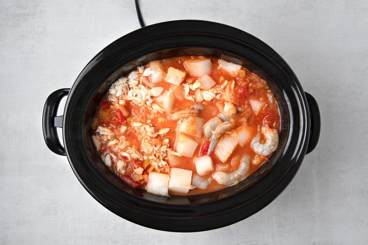 overhead shot of a black slow cooker filled with a red seafood stew; the stew contains diced potatoes, shrimp, and crab meat; the stew is set on a light gray countertop