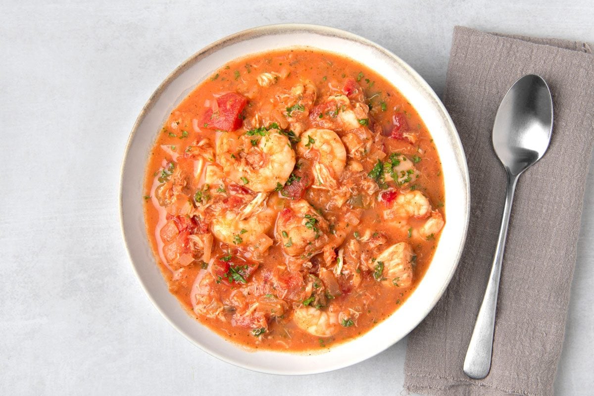 overhead shot of a bowl of seafood stew; the stew is a rich red color and contains large chunks of shrimp, fish, and vegetables; a spoon is in the bowl; the bowl is on a light gray surface with a gray napkin to the right of the bowl