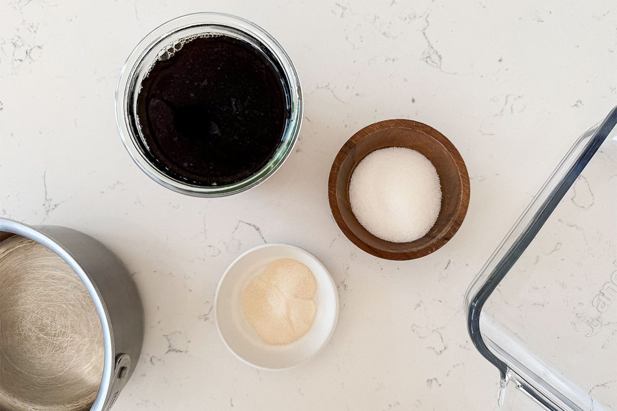 A jar of dark liquid, a small wooden bowl with white sugar, a small white bowl with a beige substance, and a metal container with a light powder arranged on a white countertop.