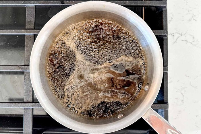 A small saucepan on a stove with liquid gently bubbling inside, appearing to be boiling coffee or a dark sauce. The stove features a black burner grate. The countertop is light-colored and speckled.