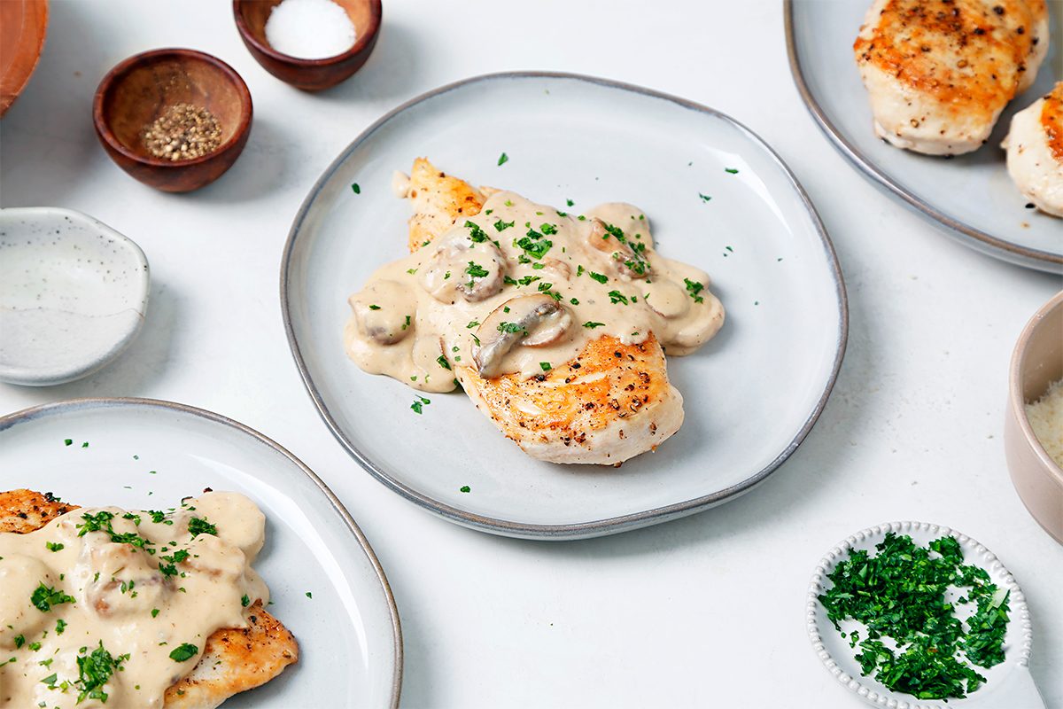 Grilled chicken topped with creamy mushroom sauce on a gray plate, garnished with chopped parsley. Surrounding the plate are small bowls of seasoning, chopped herbs, and additional grilled chicken pieces. White background.