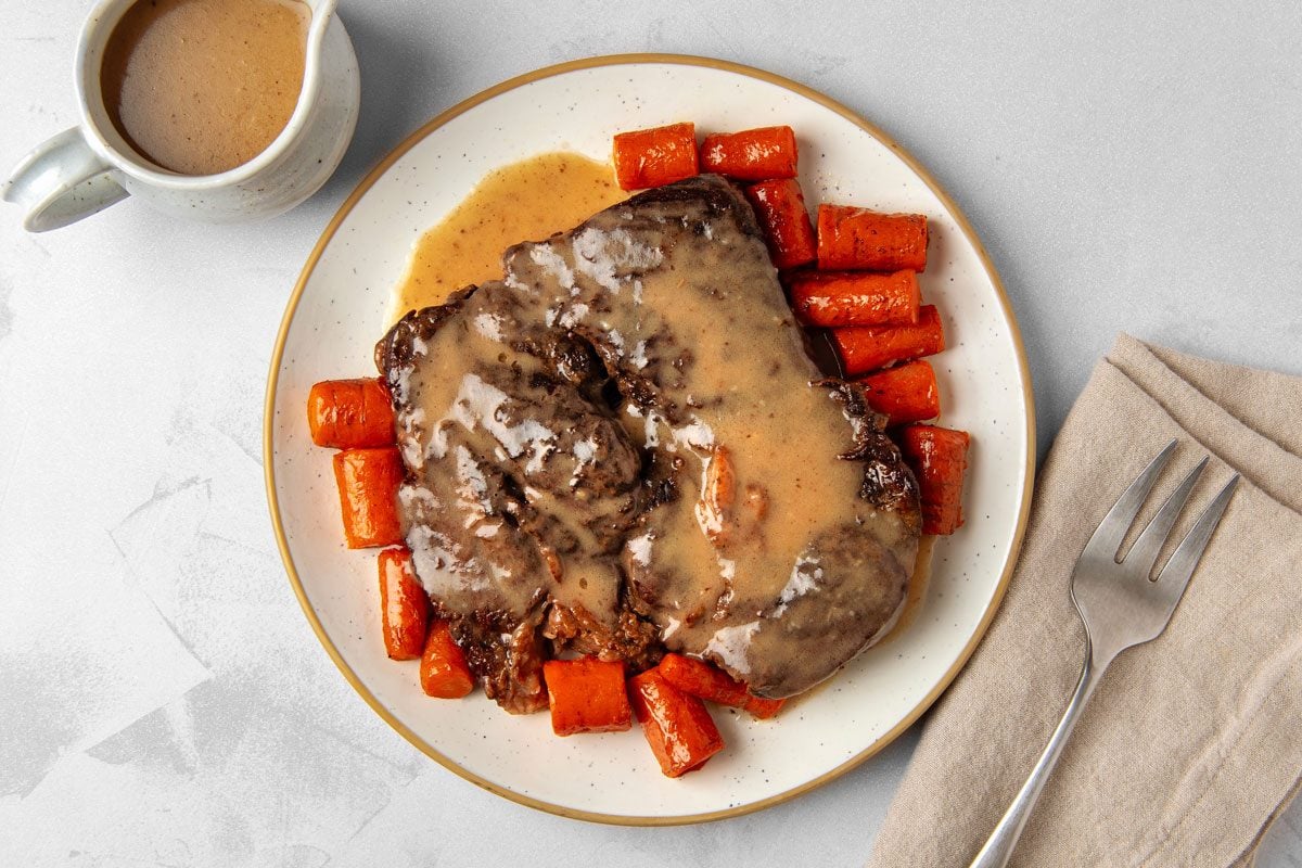 overhead shot of a plate of Dutch oven pot roast; the roast is a large cut of meat covered in a brown gravy; there is a small pitcher of gravy off to the side of the plate; a fork is placed on a tan napkin off to the side of the plate; the plate is on a white background