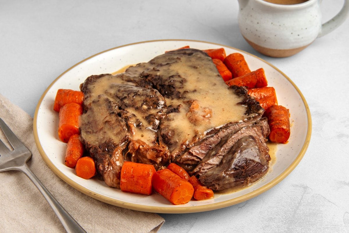 closeup shot of a plate of Dutch oven pot roast, with carrots, and gravy; a gravy boat filled with gravy sits to the left of the plate; a fork sits on a brown napkin to the right of the plate, The roast is sliced and covered in gravy; the carrots are cooked and slightly browned; the gravy boat is white with brown speckles; the napkin is a light brown with a slightly rough texture; the background is a light gray