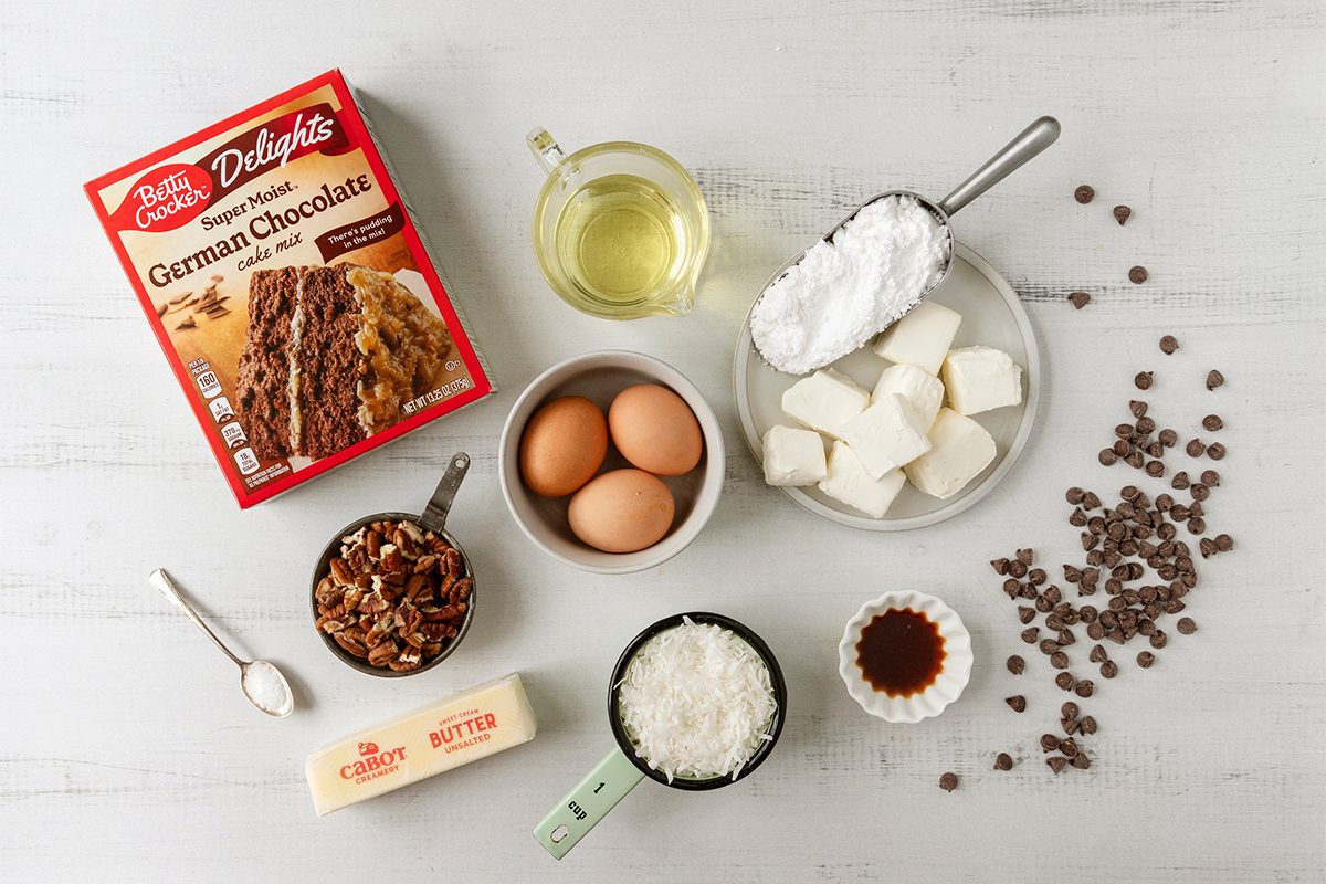 Top view of baking ingredients on a white surface: German chocolate cake mix, eggs, vegetable oil, cream cheese, powdered sugar, chocolate chips, pecans, butter, coconut flakes, and vanilla extract.