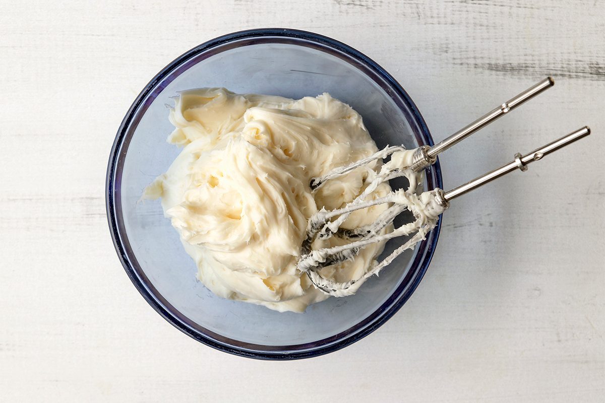 A glass bowl filled with creamy frosting or whipped topping sits on a light surface. Two metal beaters, coated with the same creamy substance, rest on the edge of the bowl.