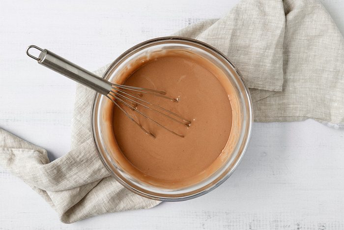 A glass bowl of light brown batter with a whisk in it, resting on a textured, light-colored cloth. The background is a white wooden surface.