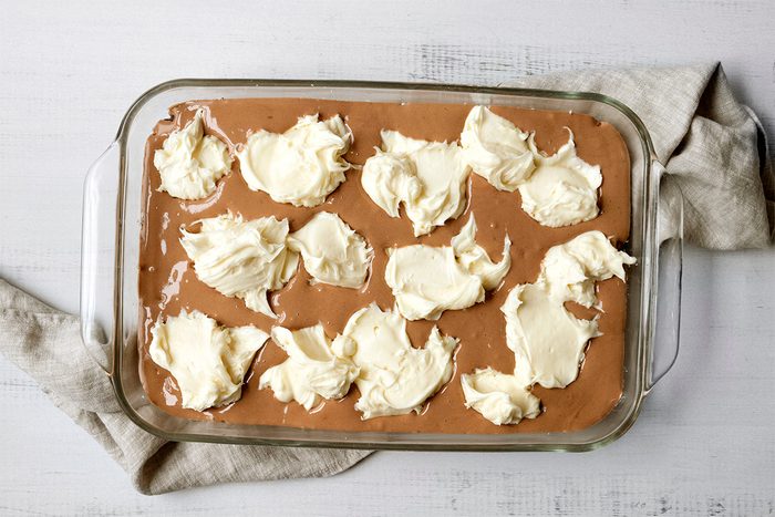 A glass baking dish filled with chocolate batter topped with dollops of white cream cheese on a light gray tablecloth.