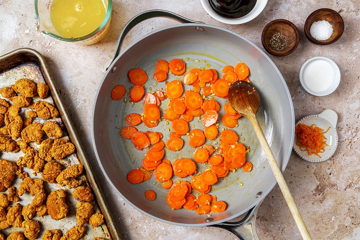A pan with sliced carrots being cooked with a wooden spoon. On the side, there are small bowls with spices, a measuring cup with liquid, and a baking sheet with breaded food pieces on a light countertop.