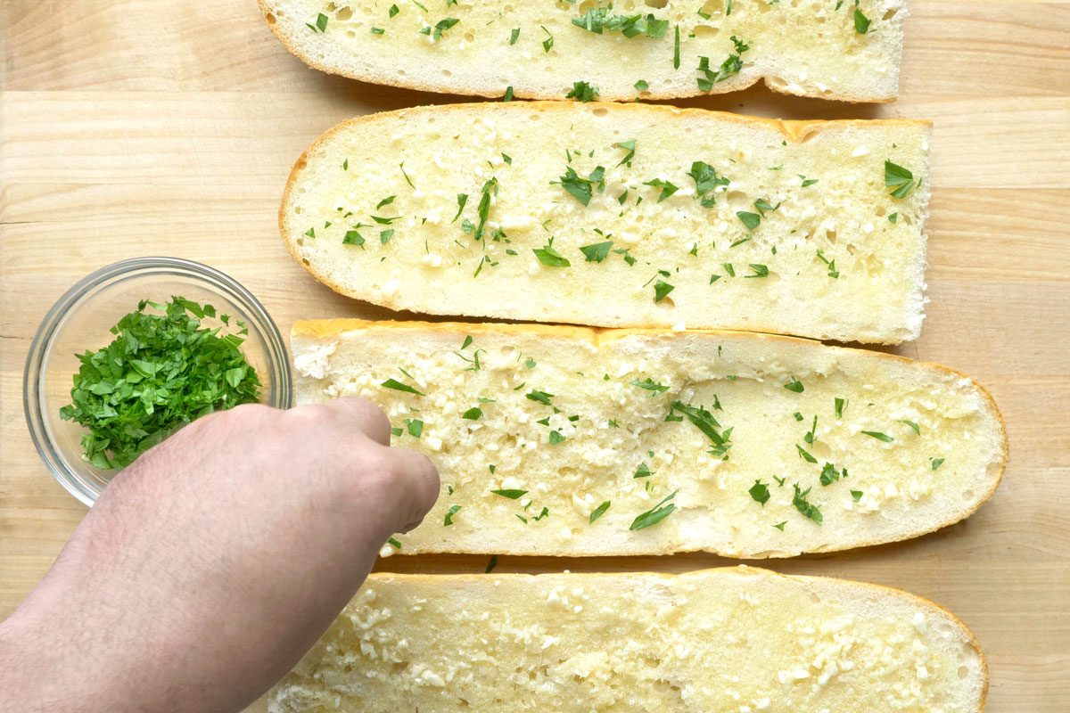 overhead shot of four slices of bread topped with garlic butter and parsley; the bread slices are arranged in a row on a wooden cutting board; a hand is holding a small glass bowl filled with chopped parsley; the hand is reaching down to sprinkle parsley on top of the bread slices