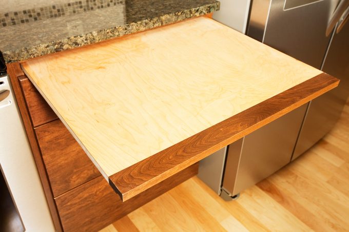 Maple wood cutting board pulls out from between a marble kitchen counter and the wooden cabinets below