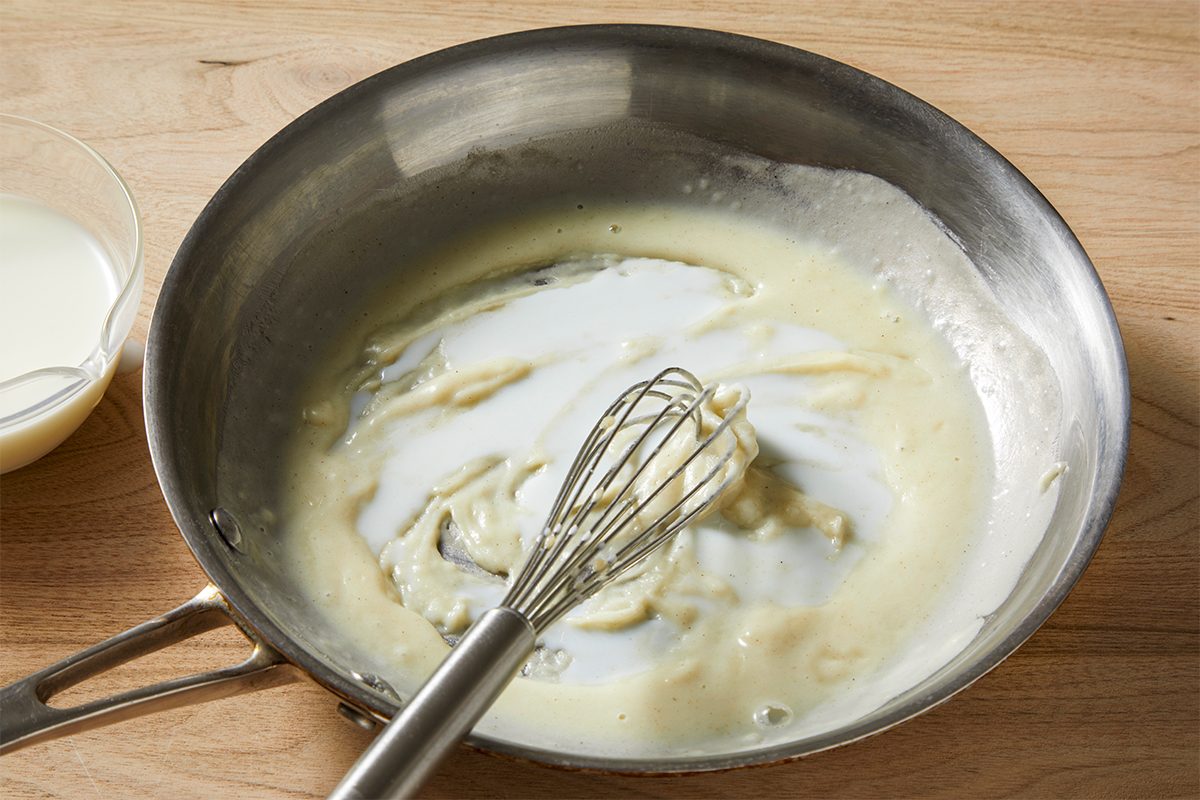 A metal whisk rests in a stainless steel pan containing a thick, creamy white sauce, partially stirred. The pan is on a wooden surface, and a small glass bowl with a white liquid is visible on the side.