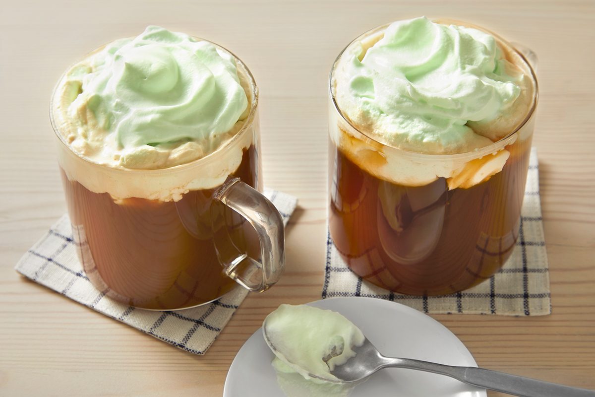 Two glass mugs of coffee topped with green-tinted whipped cream sit on checkered coasters on a wooden surface. A spoon with some whipped cream rests on a small white plate in front.