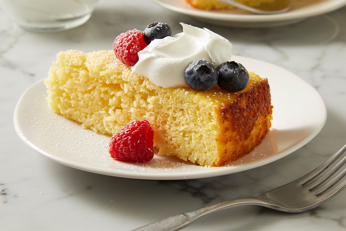 Side view of a slice of lemon ricotta cake placed on a serving plate on a white marble countertop with a fork and topped with strawberries, blueberries, and whipped cream.