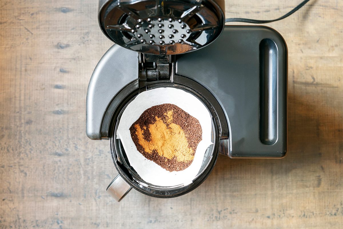 A top-down view of an open coffee maker on a wooden surface. Inside the coffee maker is a filter containing ground coffee and spices. The lid is raised, revealing the contents clearly.