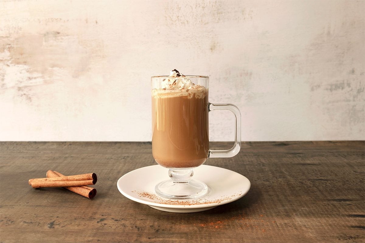 A glass mug of coffee topped with whipped cream and chocolate shavings sits on a white plate with a wooden table. Two cinnamon sticks and a small orange peel are placed on the plate beside the mug.