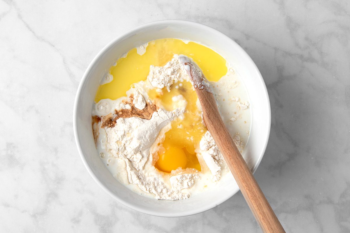 A white bowl containing ingredients for baking: flour, eggs, milk, vanilla extract, and melted butter. A wooden spoon rests in the bowl, ready to mix the ingredients, all set against a marble countertop.