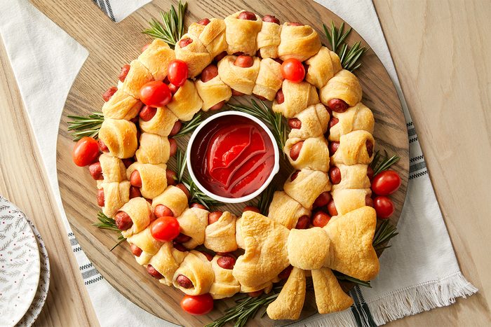 Horizontal AP Pull shot of a ring of piggies is arranged on a wooden cutting board, decorated with sprigs of rosemary and cherry tomatoes; a bowl of ketchup is placed in the center of the wreath; The cutting board sits on a white cloth with green stripes, and a stack of plates with a pattern of brown pine branches and red berries is placed on the right-hand side of the image