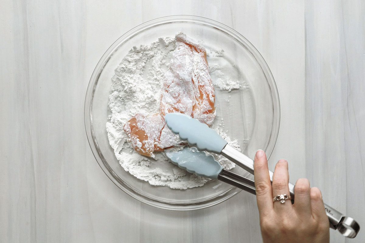 overhead shot of a person using tongs to coat a piece of chicken in flour the background is white wooden top