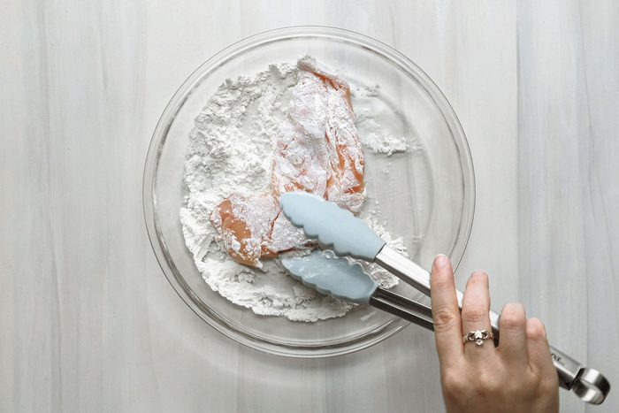 overhead shot of a person using tongs to coat a piece of chicken in flour the background is white wooden top